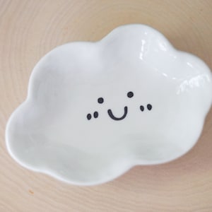 May include: A white ceramic cloud-shaped dish with a hand-painted smiley face. The dish is perfect for holding small items like jewelry or keys.