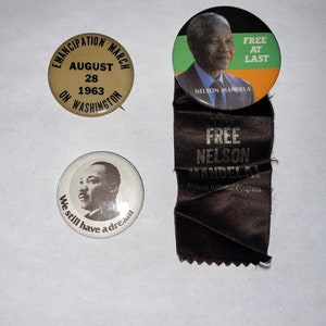May include: Three round buttons with images of Nelson Mandela, Martin Luther King Jr., and a text-only button that reads "Emancipation March August 28 1963 On Washington". The Mandela button has the text "Free at Last" and "Nelson Mandela". The King Jr. button has the text "We still have a dream". The buttons are on a white background.