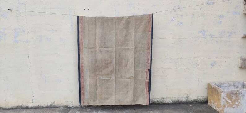 May include: A beige woven rug with a dark blue and red border hanging on a white wall.