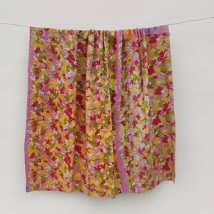 以下が含まれることがあります： ピンク、イエロー、グリーンの色合いの花柄が施されたカラフルな生地。生地は白い壁にかけられています。
