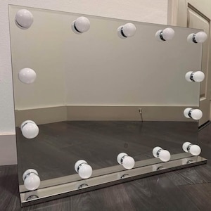 May include: A rectangular vanity mirror with a frameless glass surface and a metallic base. The mirror features twelve round white light bulbs arranged along the perimeter, providing illumination for the reflective surface.