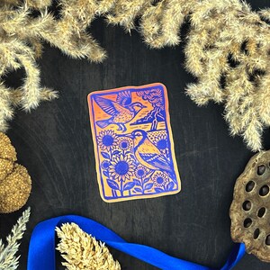 May include: A rectangular sticker with an orange border and a blue and yellow illustration of two birds, sunflowers, and a landscape. The sticker is surrounded by dried plants and a blue ribbon on a dark surface.