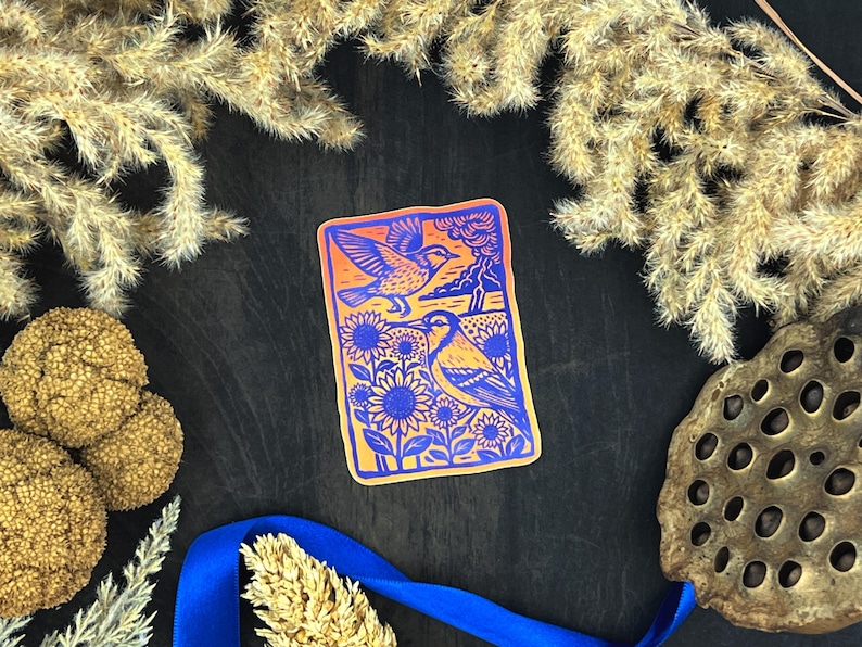 May include: A rectangular sticker with an orange border and a blue and yellow illustration of two birds, sunflowers, and a landscape. The sticker is surrounded by dried plants and a blue ribbon on a dark surface.