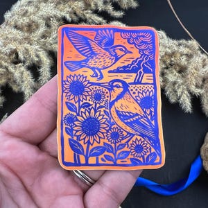 May include: A rectangular sticker with a vibrant design featuring two birds and sunflowers. The birds are depicted in blue and orange, with one in flight and the other perched among blue sunflowers. The background is orange with blue accents.