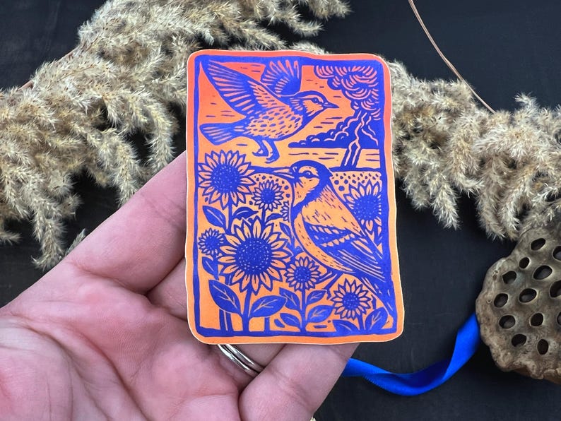 May include: A rectangular sticker with a vibrant design featuring two birds and sunflowers. The birds are depicted in blue and orange, with one in flight and the other perched among blue sunflowers. The background is orange with blue accents.