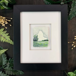 May include: A framed watercolour painting of a landscape with a cloudy sky, green trees, and a field of grass.