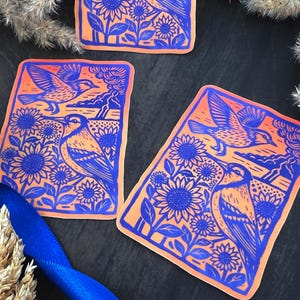 May include: Rectangular stickers with a blue and orange design. The stickers feature a bird, sunflowers, and a landscape. The bird is depicted in flight and perched among the sunflowers. The background is orange, and the design is blue.
