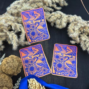 May include: Three rectangular stickers with a bird and sunflower design. The stickers feature a blue and orange color scheme. The design includes a bird in flight, sunflowers, and a landscape. The stickers are arranged on a dark surface with dried plants and a blue ribbon.