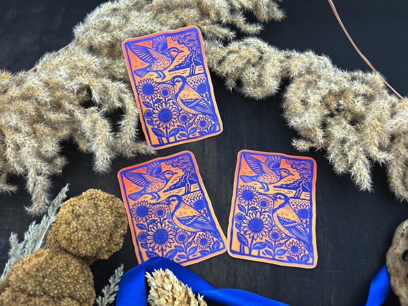 May include: Three rectangular stickers with a bird and sunflower design. The stickers feature a blue and orange color scheme. The design includes a bird in flight, sunflowers, and a landscape. The stickers are arranged on a dark surface with dried plants and a blue ribbon.
