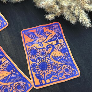 May include: Orange and blue sticker featuring a linocut design of two birds, sunflowers, and a landscape. The birds are depicted in flight and perched among the sunflowers. The sticker has a rectangular shape.