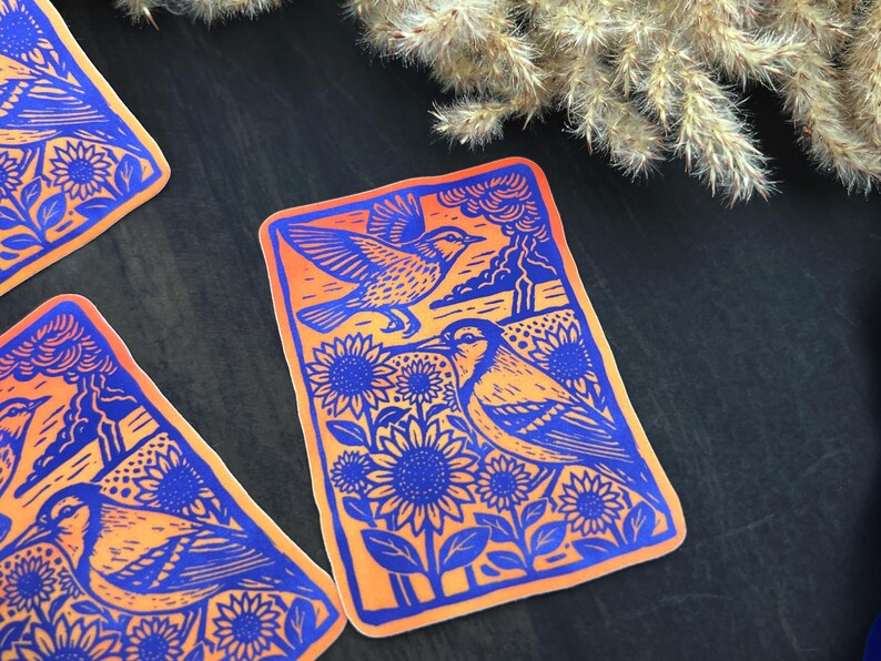 May include: Orange and blue sticker featuring a linocut design of two birds, sunflowers, and a landscape. The birds are depicted in flight and perched among the sunflowers. The sticker has a rectangular shape.