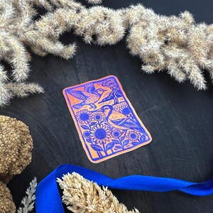 May include: A rectangular sticker with a vibrant design featuring two birds, sunflowers, and a landscape in shades of orange and blue. The sticker is surrounded by dried plants and a blue ribbon.