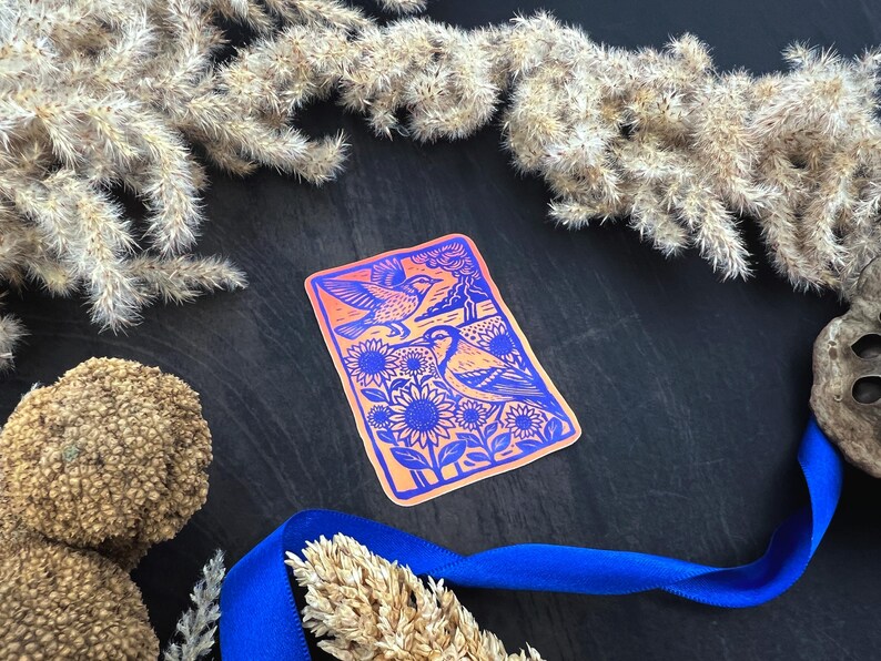May include: A rectangular sticker with a vibrant design featuring two birds, sunflowers, and a landscape in shades of orange and blue. The sticker is surrounded by dried plants and a blue ribbon.