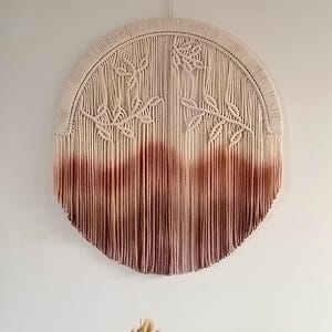 Peut inclure: Une tenture murale ronde en macramé avec un motif floral. La partie supérieure est de couleur crème naturelle, passant à un bordeaux profond en bas. Le design comprend des motifs détaillés de feuilles et de fleurs.