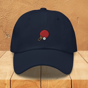 May include: A navy blue baseball cap with an embroidered red and brown table tennis bat and white ball.