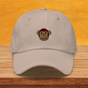 Può includere: Un cappello da baseball beige con un viso di scimmia ricamato. La scimmia ha un viso marrone, orecchie rosse e un sorriso giallo.