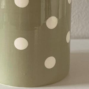 May include: A close-up of a ceramic vase with a matte finish. The vase is olive green with evenly spaced white polka dots. The vase is cylindrical and has a smooth surface.