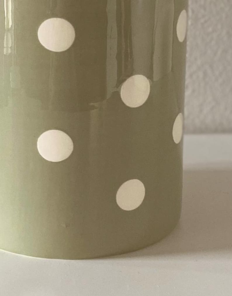 May include: A close-up of a ceramic vase with a matte finish. The vase is olive green with evenly spaced white polka dots. The vase is cylindrical and has a smooth surface.
