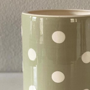 May include: A close-up of a ceramic mug with a light green exterior and white polka dots. The interior of the mug is a light beige color. The mug has a glossy finish and reflects light.