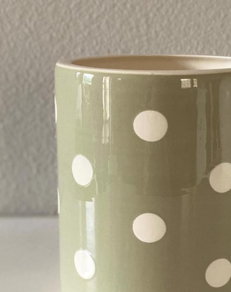 May include: A close-up of a ceramic mug with a light green exterior and white polka dots. The interior of the mug is a light beige color. The mug has a glossy finish and reflects light.