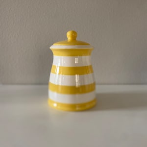 May include: A ceramic canister with a glossy finish featuring horizontal yellow and white stripes. The container has a slightly curved body and a matching removable lid with a small, rounded knob handle at the very top center.