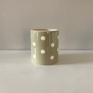 May include: A cylindrical ceramic planter in a muted green color, adorned with evenly spaced white polka dots. The planter has a smooth, glossy finish and a neutral interior. It sits on a white surface against a light gray background.