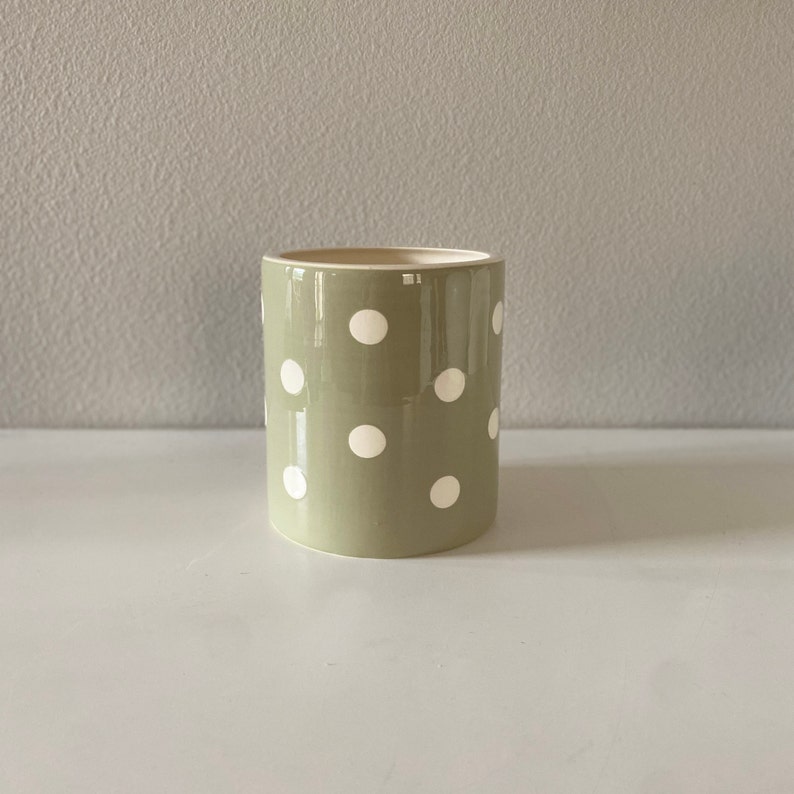 May include: A cylindrical ceramic planter in a muted green color, adorned with evenly spaced white polka dots. The planter has a smooth, glossy finish and a neutral interior. It sits on a white surface against a light gray background.
