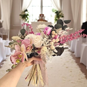 May include: A wedding bouquet with white roses, pink orchids, and dried flowers, held by a hand. The bouquet is tied with a pale pink ribbon. The background shows a wedding venue with white curtains and a decorated aisle.