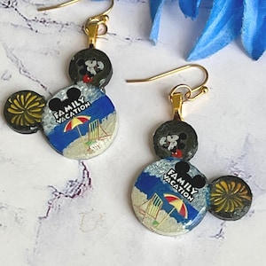 May include: Gold-tone dangle earrings featuring a Mickey Mouse design. The earrings have a black head and ears, with a circular beach scene that reads "Family Vacation". The earrings are a fun accessory.