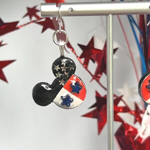 May include: A dangle earring shaped like a mouse head silhouette. The design features a black top section with silver star accents, and a lower section divided into red and white panels, each decorated with a small, textured blue star.