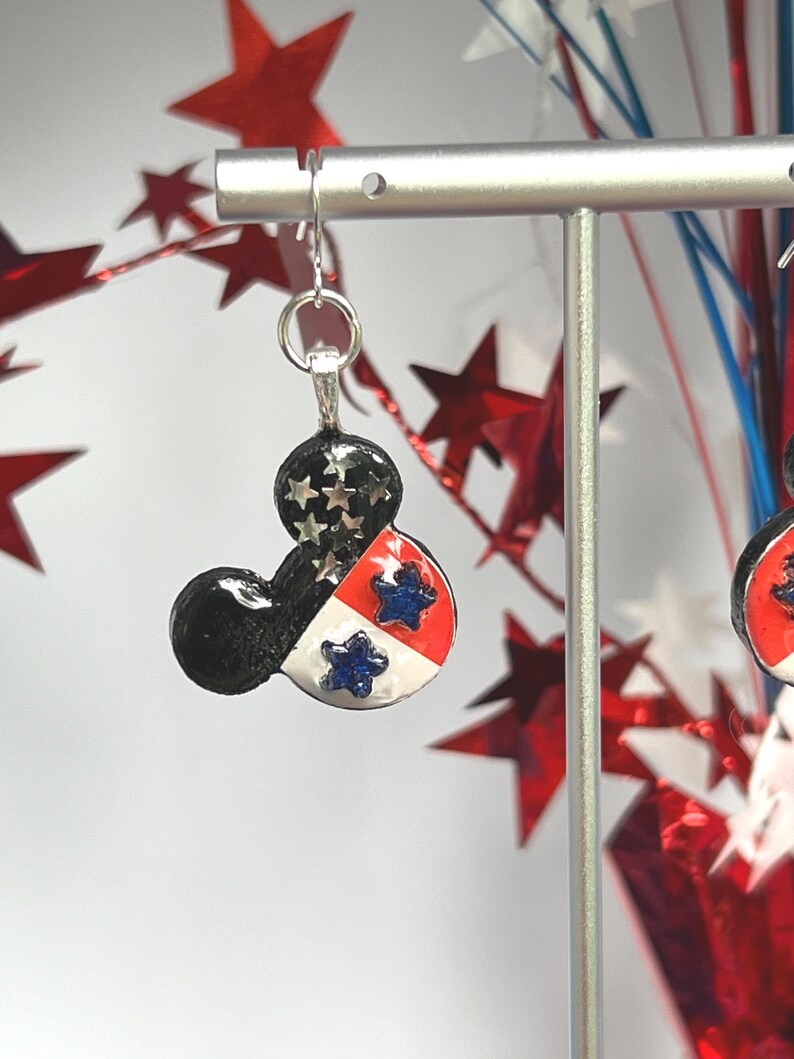 May include: A dangle earring shaped like a mouse head silhouette. The design features a black top section with silver star accents, and a lower section divided into red and white panels, each decorated with a small, textured blue star.