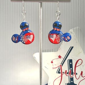 May include: A pair of dangle earrings featuring a mouse-shaped silhouette in red, white, and blue. The design includes a heart with an American flag pattern, firework graphics, and striped accents on the ears, suspended from silver-toned hooks.