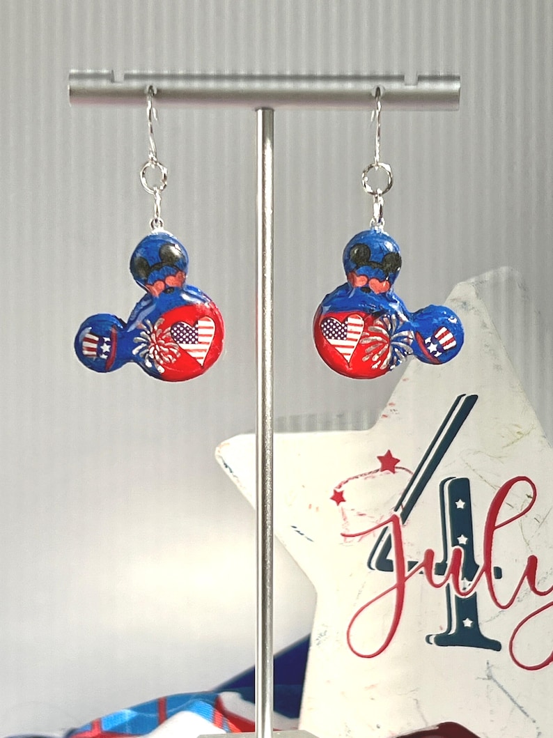 May include: A pair of dangle earrings featuring a mouse-shaped silhouette in red, white, and blue. The design includes a heart with an American flag pattern, firework graphics, and striped accents on the ears, suspended from silver-toned hooks.