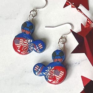 May include: A pair of dangle earrings featuring a blue, red, and white mouse-shaped charm. The design includes a heart with an American flag pattern, a firework burst, and a small top hat with red and white stripes and blue stars on a silver hook.