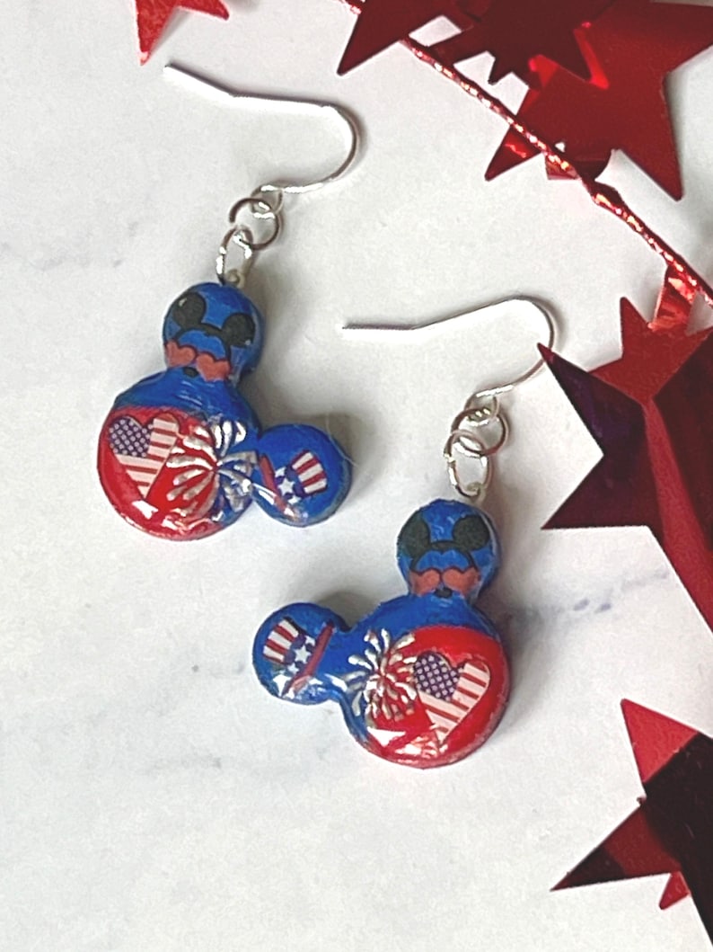 May include: A pair of dangle earrings featuring a blue, red, and white mouse-shaped charm. The design includes a heart with an American flag pattern, a firework burst, and a small top hat with red and white stripes and blue stars on a silver hook.