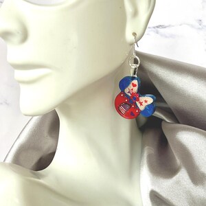 May include: A dangling earring featuring a mouse-ear silhouette painted in red, white, and blue. The design includes a central bow with star and stripe patterns, small red hearts, and a silver-toned metal hook for attachment to the ear.