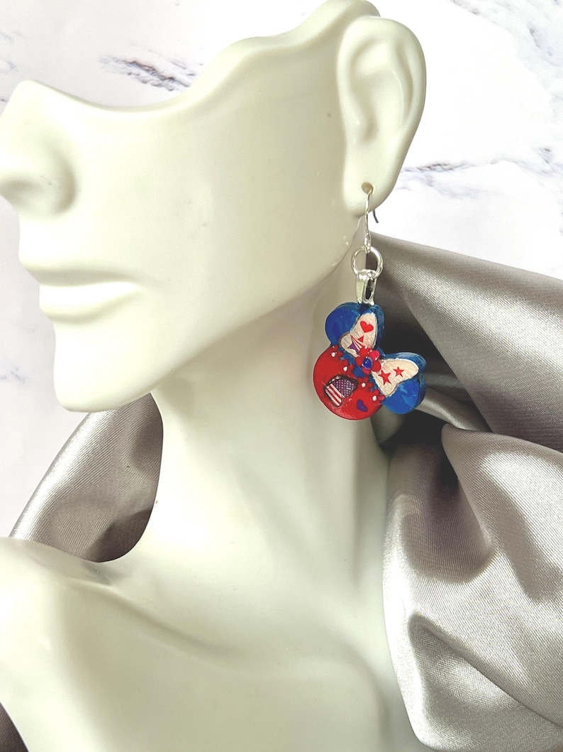May include: A dangling earring featuring a mouse-ear silhouette painted in red, white, and blue. The design includes a central bow with star and stripe patterns, small red hearts, and a silver-toned metal hook for attachment to the ear.