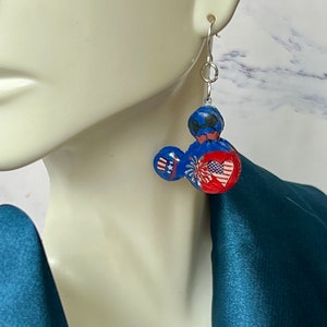 May include: A dangle earring featuring a blue, red, and white mouse-shaped charm. The charm displays a patriotic theme with a small top hat, a firework burst, and a heart containing a star-spangled banner pattern, suspended from a silver hook.