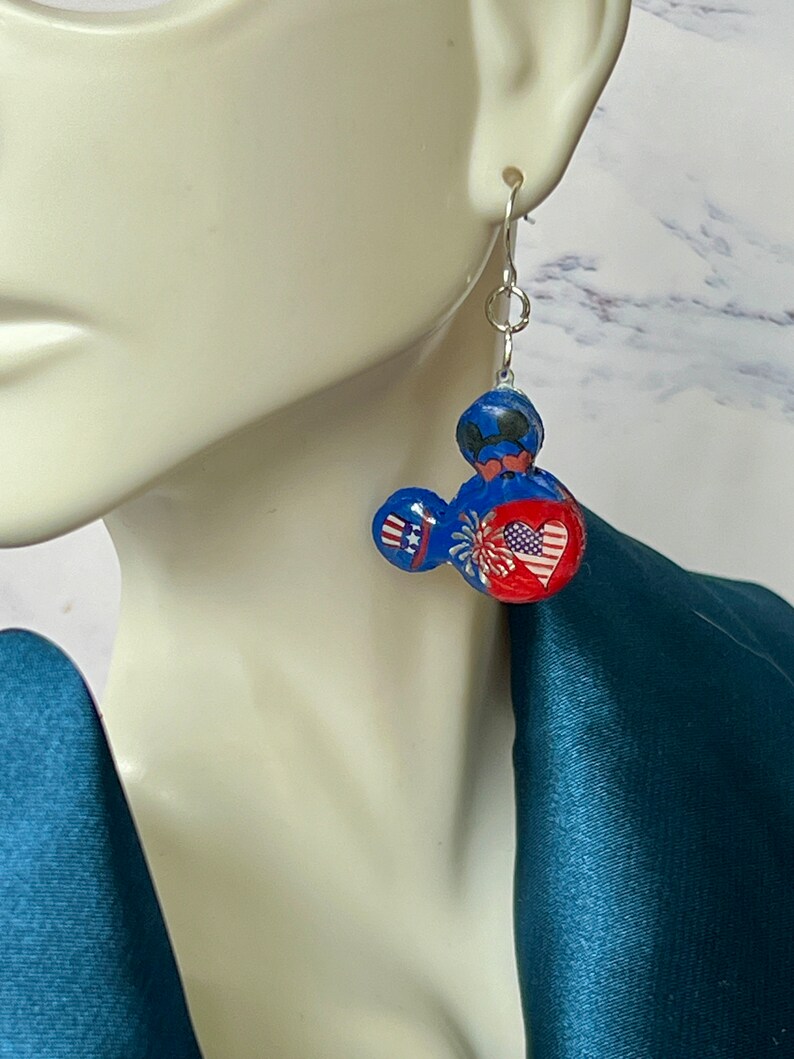 May include: A dangle earring featuring a blue, red, and white mouse-shaped charm. The charm displays a patriotic theme with a small top hat, a firework burst, and a heart containing a star-spangled banner pattern, suspended from a silver hook.