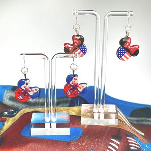 May include: Four dangle earrings shaped like mouse ears, displayed on two clear acrylic stands. Each earring features a patriotic design with red, white, and blue colors, incorporating star and stripe patterns. The earrings hang from silver-toned hooks.