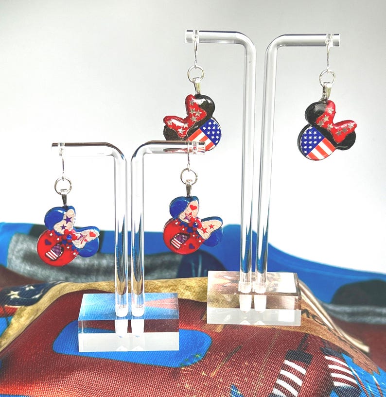 May include: Four dangle earrings shaped like mouse ears, displayed on two clear acrylic stands. Each earring features a patriotic design with red, white, and blue colors, incorporating star and stripe patterns. The earrings hang from silver-toned hooks.