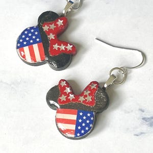 May include: A pair of dangle earrings featuring a mouse-shaped silhouette with a black base. The design includes a red bow adorned with silver stars and a face section patterned with the American flag, featuring red and white stripes and blue stars.