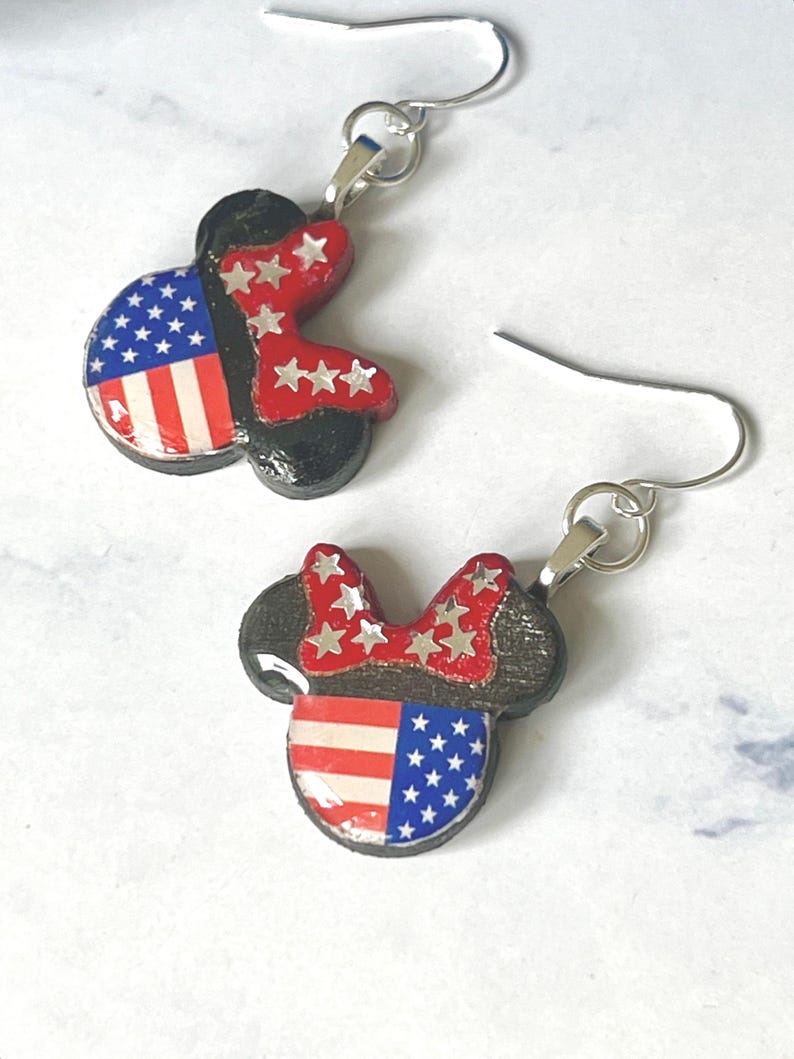 May include: A pair of dangle earrings featuring a mouse-shaped silhouette with a black base. The design includes a red bow adorned with silver stars and a face section patterned with the American flag, featuring red and white stripes and blue stars.