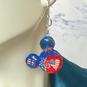 May include: A dangle earring featuring a three-circle mouse-shaped charm in blue and red. The design includes a small top hat with red and white stripes, a white firework burst, and a heart with a red, white, and blue American flag pattern.