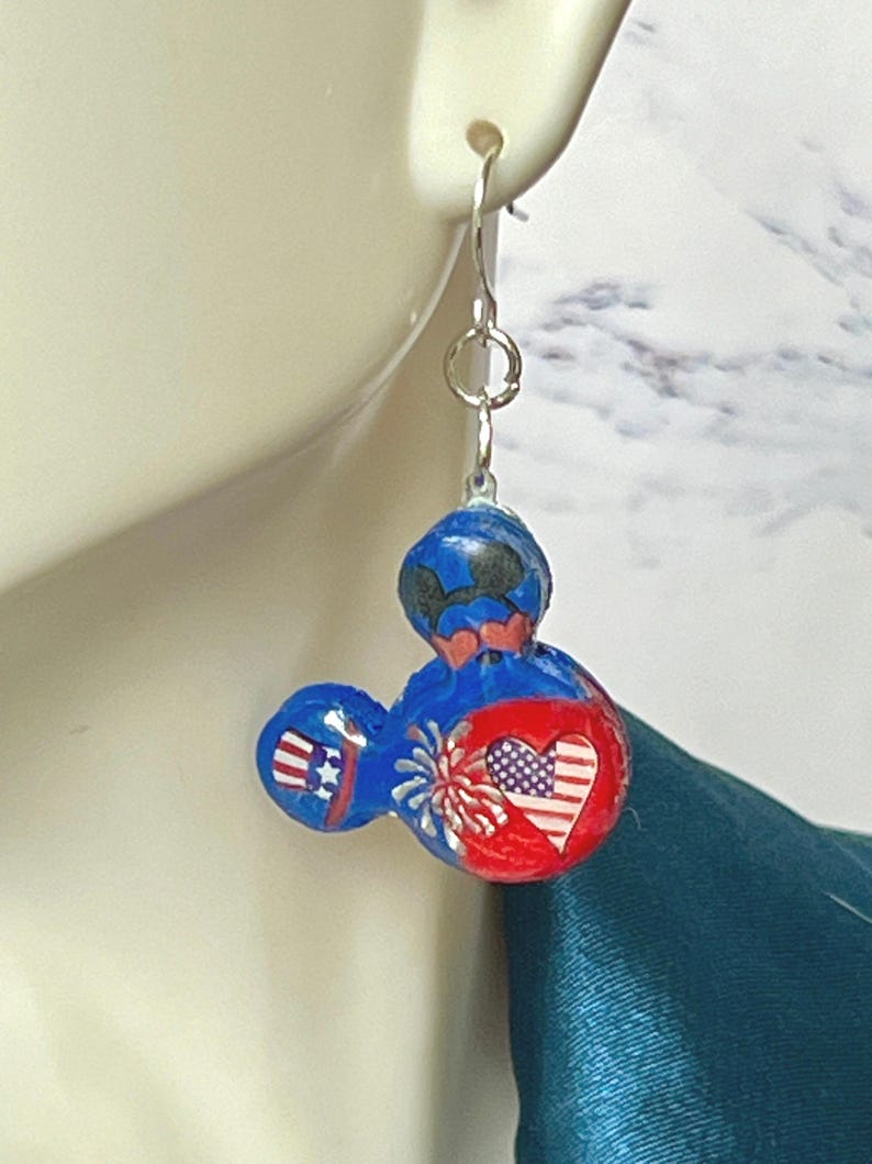 May include: A dangle earring featuring a three-circle mouse-shaped charm in blue and red. The design includes a small top hat with red and white stripes, a white firework burst, and a heart with a red, white, and blue American flag pattern.