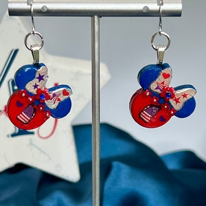 May include: A pair of dangle earrings featuring a mouse-ear silhouette in red, white, and blue. The design includes silver accents, small red flowers, blue rhinestones, and star and heart motifs. Each earring hangs from a silver-toned metal hook.