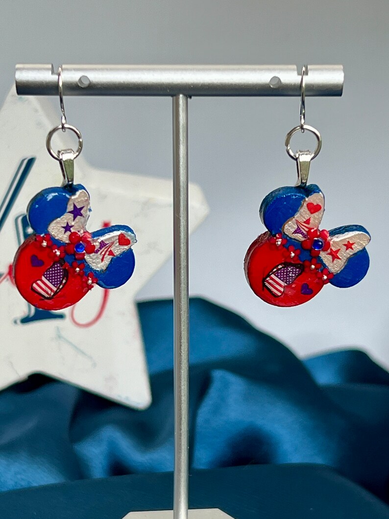 May include: A pair of dangle earrings featuring a mouse-ear silhouette in red, white, and blue. The design includes silver accents, small red flowers, blue rhinestones, and star and heart motifs. Each earring hangs from a silver-toned metal hook.