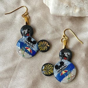 May include: A pair of gold-toned dangle earrings. Each earring features a black Mickey Mouse head, a blue and white beach scene with the text "FAMILY VACATION", and a yellow burst accent. The earrings are designed for pierced ears.