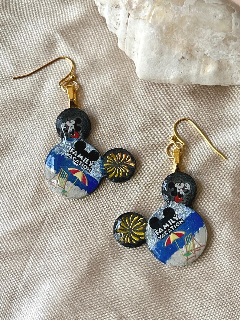 May include: A pair of gold-toned dangle earrings. Each earring features a black Mickey Mouse head, a blue and white beach scene with the text "FAMILY VACATION", and a yellow burst accent. The earrings are designed for pierced ears.
