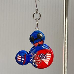 May include: A dangling earring featuring a blue, red, and white mouse-shaped charm. The charm displays a patriotic theme with a heart containing an American flag pattern, a firework burst, and a small top hat with stars and stripes on the ear.